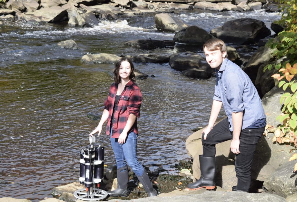 Two individuals standing on the shore of a river with a submersible device
