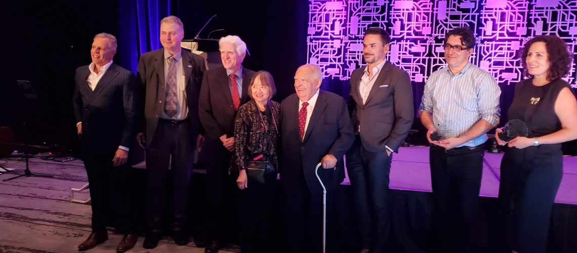 A group of eight people are standing in a line on a stage with a purple geometric patterned backdrop. The individuals are dressed in formal attire, including suits and dresses. One person is holding a cane. The stage has dark curtains and the floor has a patterned carpet.