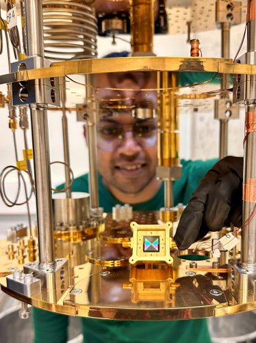 Individual in green shirt and black gloves operating a scientific instrument composed of metallic rods, wires, and circular plates. A small square device with a colorful display is centered in the apparatus.