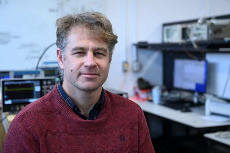 Person seated in a lab, wearing a red sweater over a dark shirt. Background includes computer monitors, electronic devices, and cables on a desk.