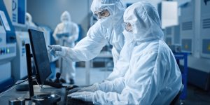 Factory Cleanroom: Engineer and Scientist Wearing Coveralls and Masks Have Discussion, Use Computer Showing Infrastructure System Control. In Background CNC Machinery, Electronics Equipment