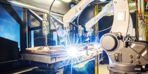 A high tech robotic welder is in use, creating a starburst of bright white light at the contact point.  The machine's large, segmented arm extends outward and down to the workspace in front of it.  Fluorescent light glows from ceiling above.